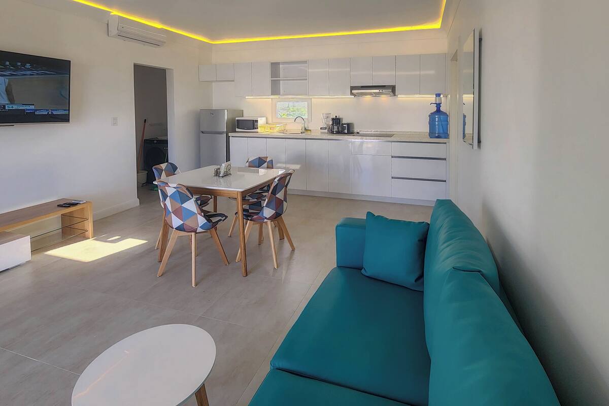 Een moderne woonkamer met een blauwgroene bank, een eettafel met gestreept stoelen, een aan de muur bevestigde televisie en een keuken met witte kasten en een magnetron.