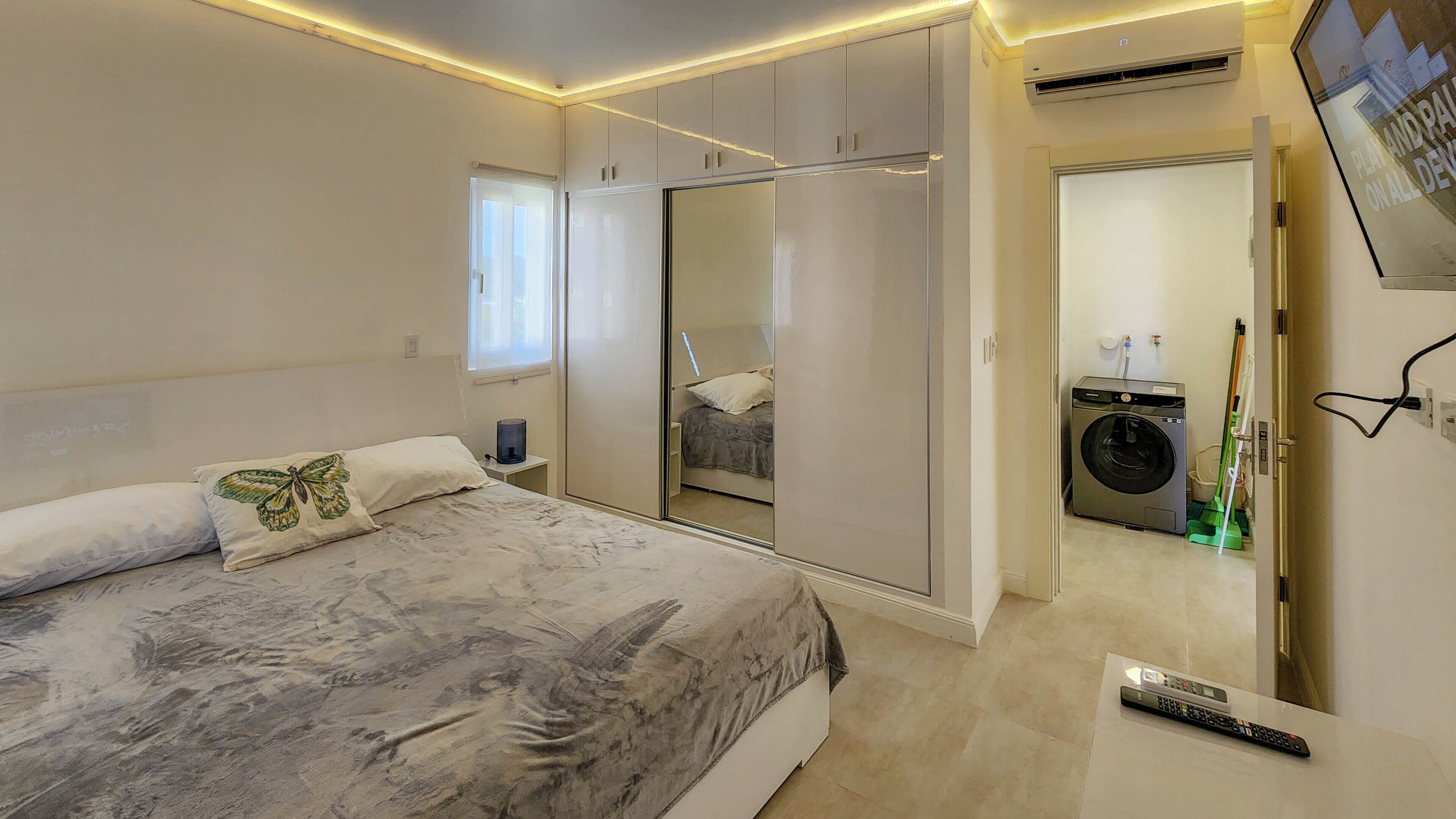 Un dormitorio con cama, armario, lavadora y televisor montado en la pared.