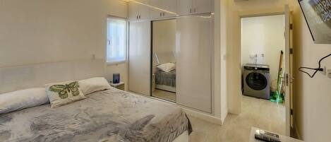Un dormitorio con cama, armario, lavadora y televisor montado en la pared.