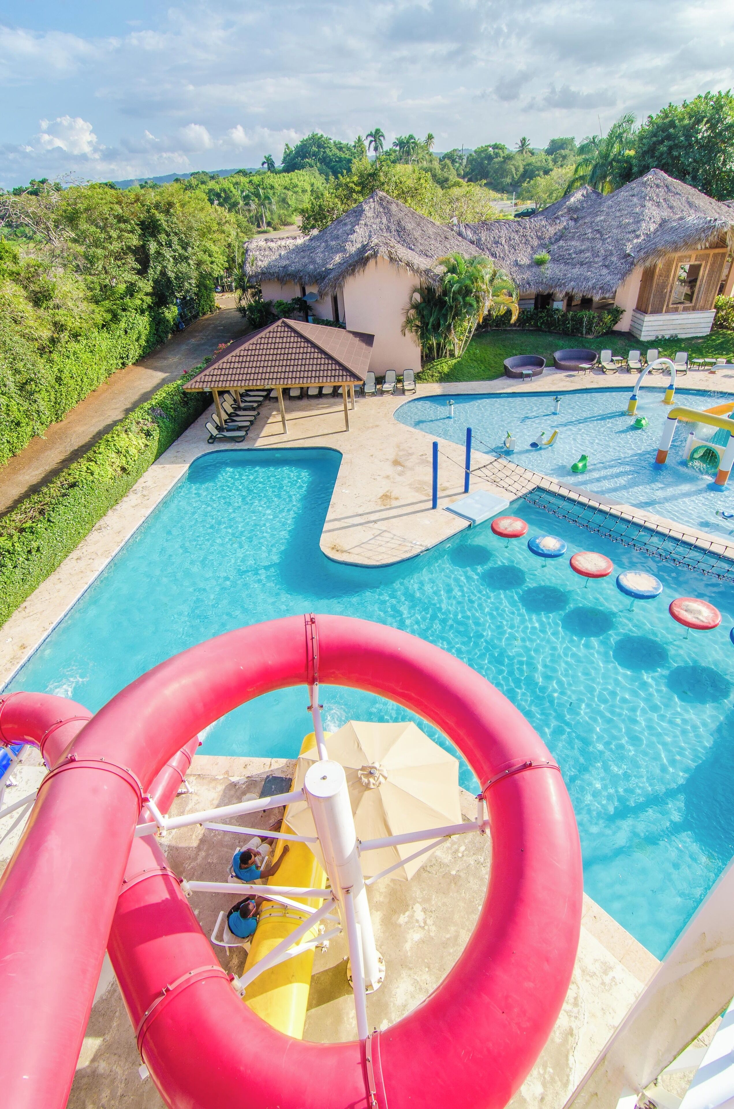 A water park with a large pool, a red water slide, and thatched-roof buildings.