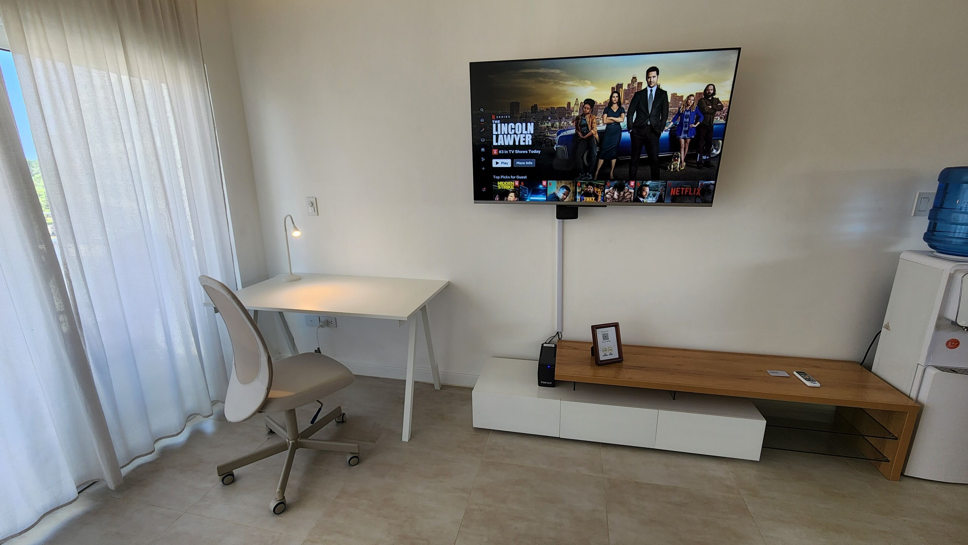 A modern hotel room with a flat-screen TV mounted on the wall, a white desk with a chair, a wooden bench, and a small table with a remote control.
