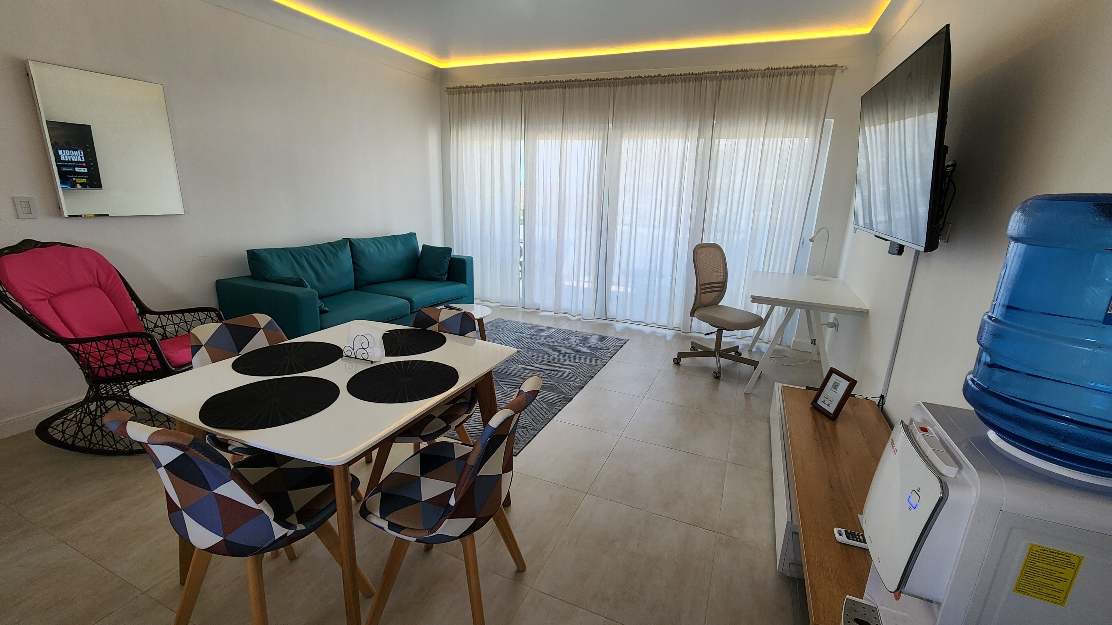 Un salon moderne avec un canapé de couleur turquoise, une table à manger avec des chaises à motifs, un fauteuil rouge, un téléviseur fixé au mur et un grand distributeur d’eau.