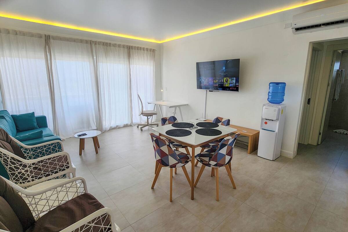 Een moderne woonkamer met een bank, een eettafel met stoelen, een aan de muur bevestigde televisie en een waterdispenser.