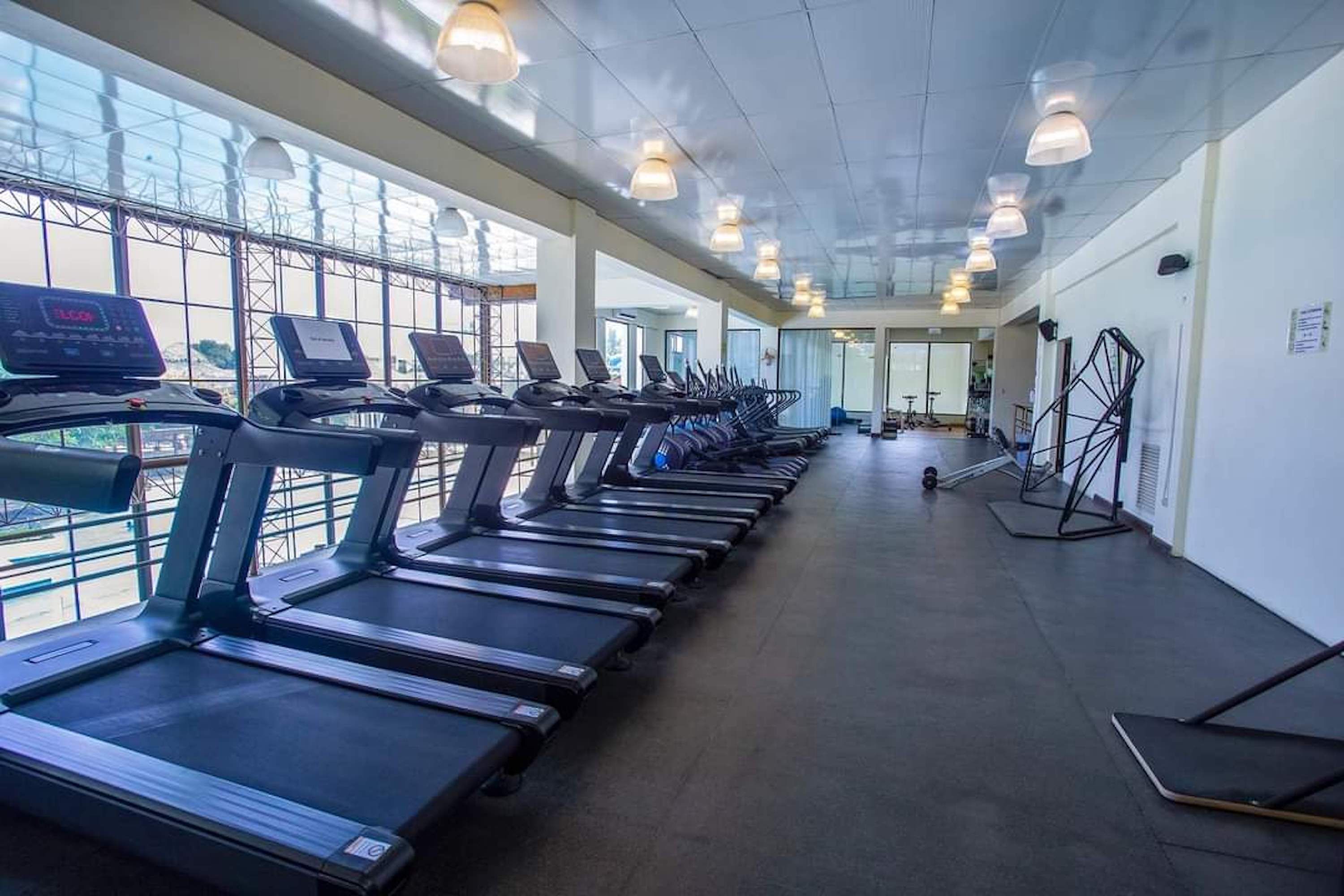 A gym with treadmills aligned along the walls, large windows, and ceiling lights.