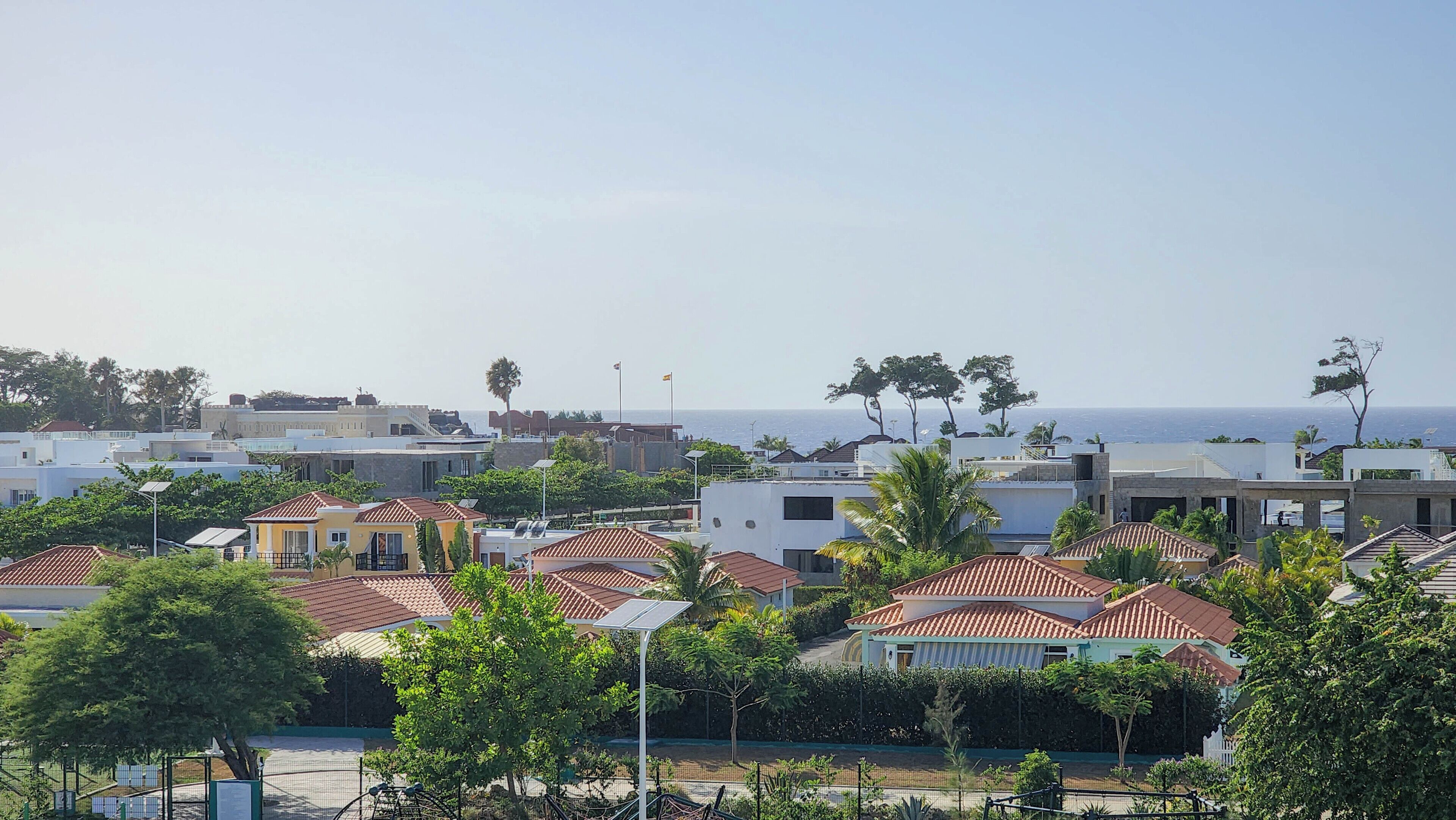 A coastal residential area with houses featuring red-tiled roofs, surrounded by greenery and palm trees.