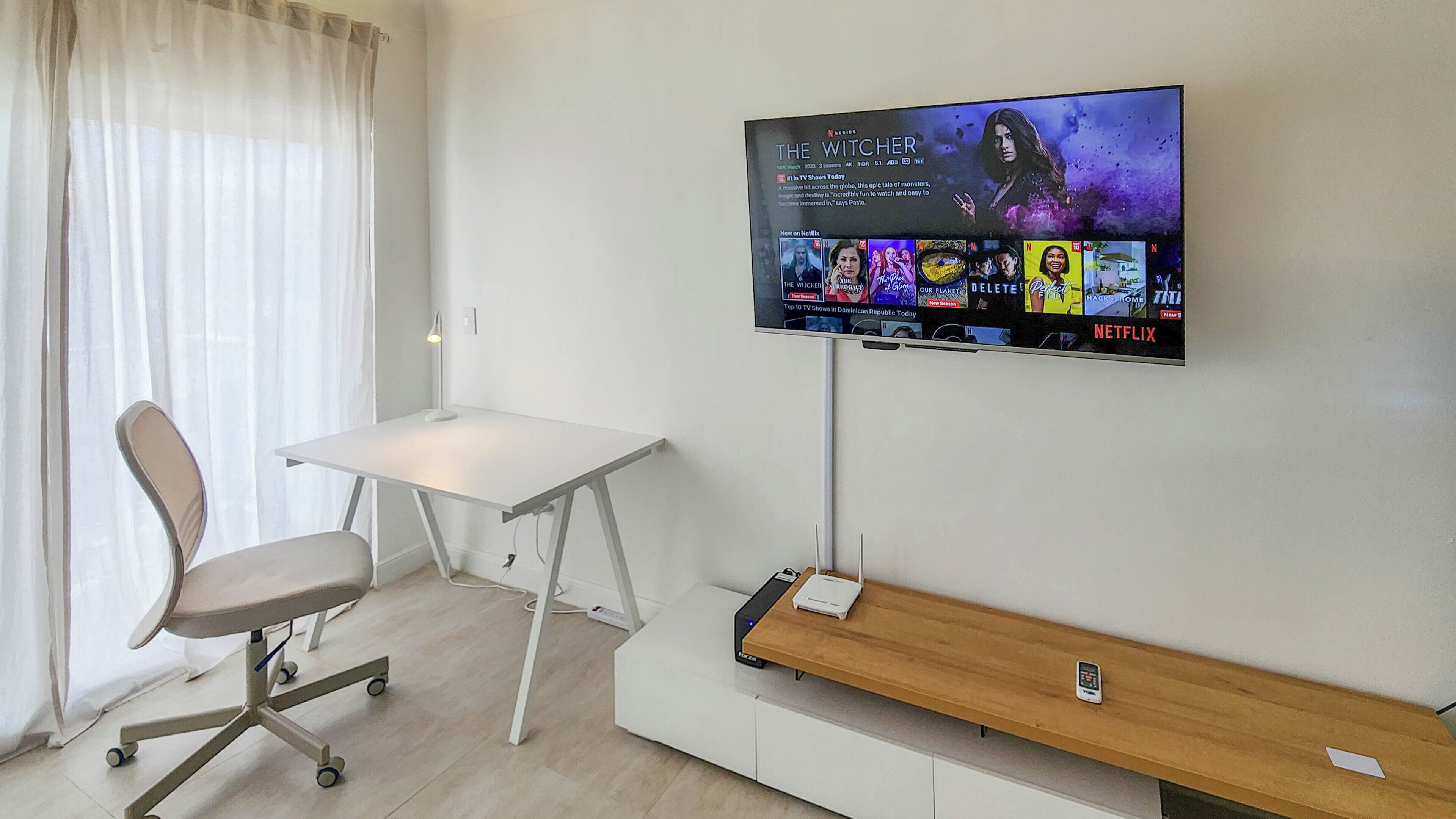Moderni huone, jossa on seinälle asennettu taulutelevisio, jossa näkyy Netflixin käyttöliittymä, valkoinen työpöytä, tuoli ja puinen penkki.