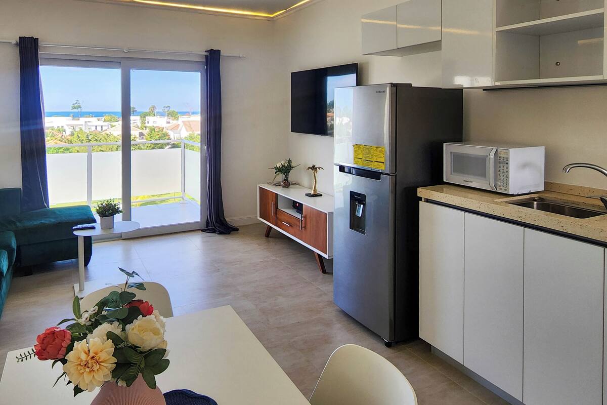 Een moderne keuken met een inbouwmagnetron, koelkast en een klein spoelbakje. Er staat een eettafel met stoelen en een bloevas. In de woonkamer is een bank en een klein tafeltje met een plantje te zien.