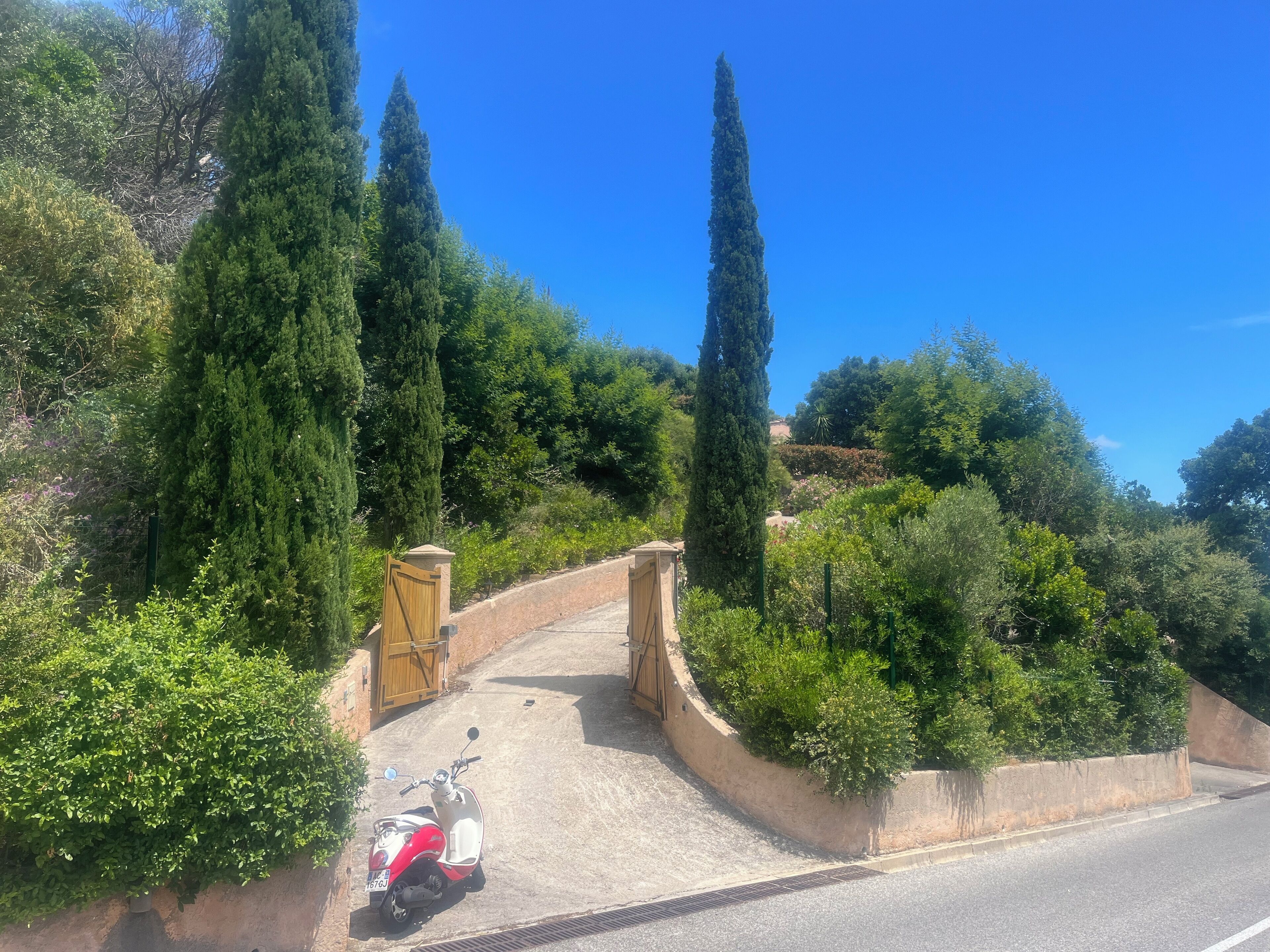 Un chemin pavé mène à une entrée fermée par un portail, qui est elle-même entourée d’une végétation luxuriante et de grands arbres.