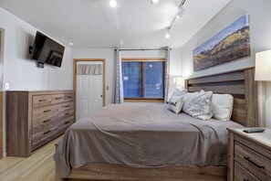 A bedroom with a bed, a TV mounted on the wall, a window with curtains, a painting on the wall, and a bedside table with a lamp. - Modern condo, Looking up to Mount Royal, Main Street Location 107 (Frisco)