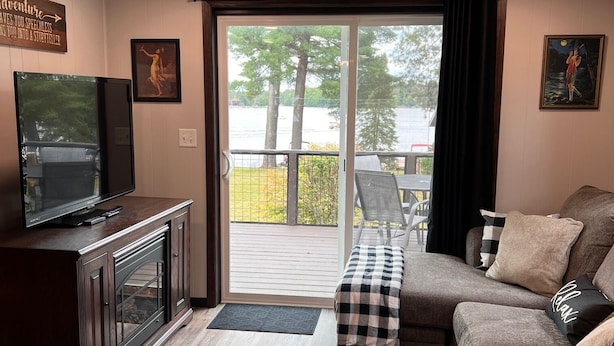 Ett vardagsrum med en tv-apparat på ett skåp, en soffa med kuddar, utsikt över en terrass och träd, samt en tavla på väggen.
