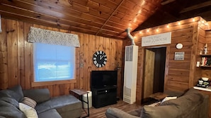 Interior acollidor d'una cabana de fusta amb una llar de foc, parets de fusta i un rellotge gran.