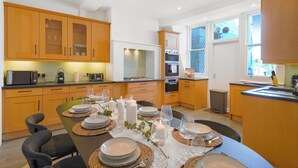 A modern kitchen with wooden cabinets, a dining table set for six with plates, glasses, and candles, and a view of the outside through large windows.