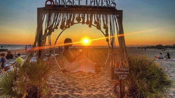 Eine Strandansicht mit einem Pavillon mit dem Schriftzug „Sunset Lovers“ und einem Schild, auf dem steht: „Dieser Moment ereignet sich einmal im Leben.“