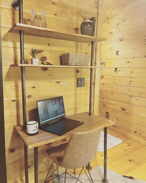 A wooden cabin interior with a desk, laptop, coffee mug, shelves with decorative items, and a chair.