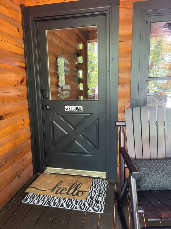 Entrada de una cabaña de madera con una puerta negra que dice “WELCOME” y un felpudo que dice “hello”.