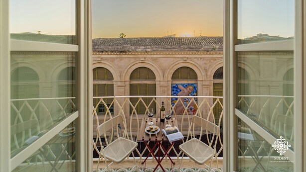 A balcony with two wicker chairs, a table with a bottle of wine, glasses, and a menu. In the background, there is a building with arches and a mural. - La Terrazza sull'Antico Mercato (Syracuse)