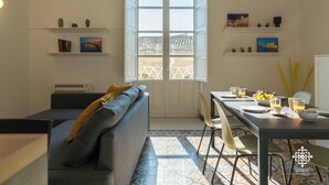 A modern living room with a sofa, a dining table set for two, and a large window with a view of a building. - La Terrazza sull'Antico Mercato (Syracuse)