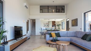Ein großzügiger Wohnbereich mit einer großen Couch, einem Couchtisch und einem Flachbildfernseher. Eine Treppe führt auf eine obere Ebene, und im Hintergrund ist eine Küche erkennbar.