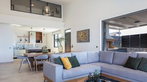 Ein modernes Wohnzimmer mit einem grauen Sofa, einem Esstisch und Stühlen. Im Hintergrund befindet sich eine Küche mit weißen Schränken und einem großen Fenster.