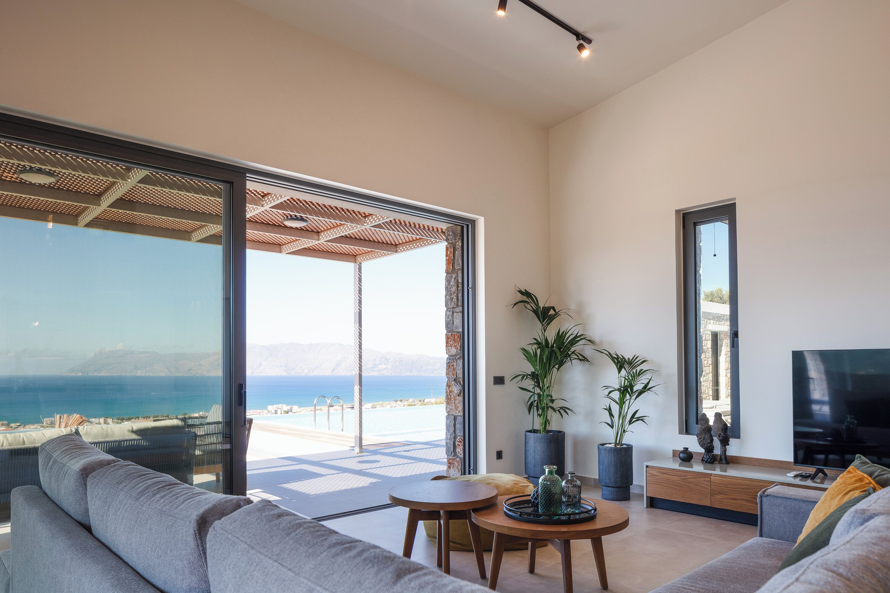 A modern living room with a large sliding glass door leading to a balcony, a flat-screen TV, and a seating area with a sofa and a small round table.