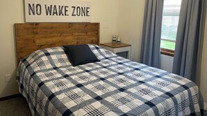 A bedroom with a bed, a bedside table, a "NO WAKE ZONE" sign, and a window with curtains.