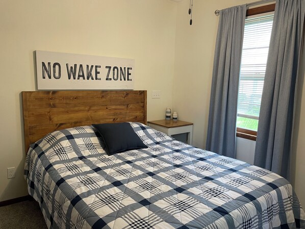 Un dormitorio con cama, mesita de noche, un letrero que dice "NO WAKE ZONE" y una ventana con cortinas.