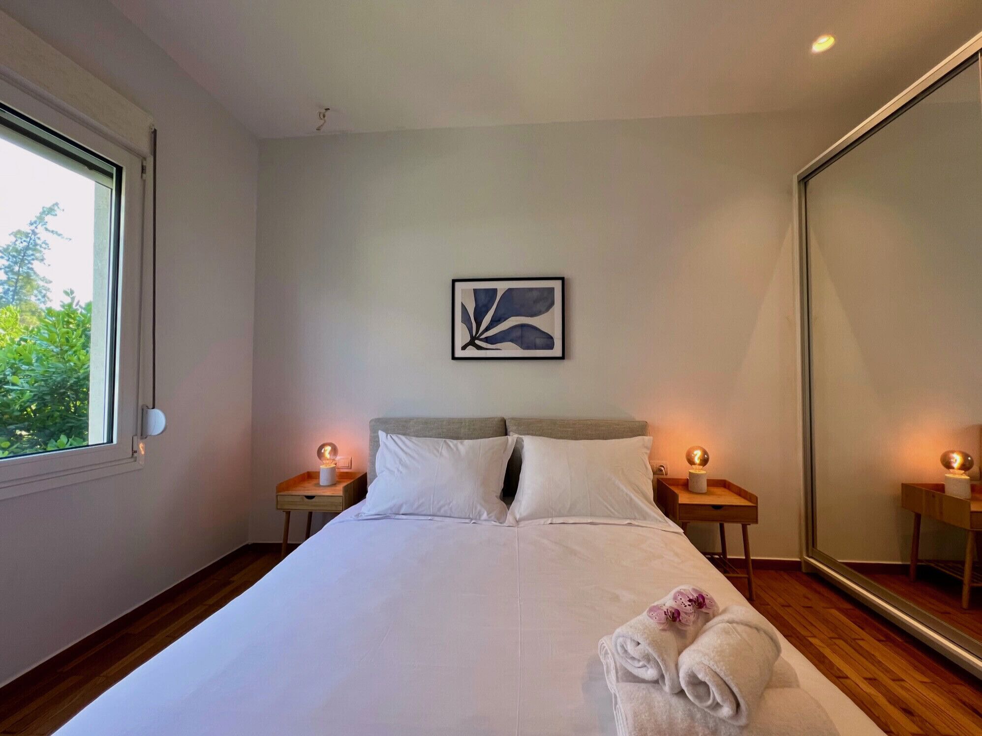 Une chambre à coucher moderne avec un grand lit, deux tables de chevet avec des lampes, une œuvre d’art abstraite encadrée et un grand miroir.