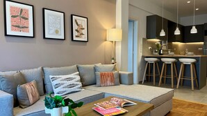 A modern living room with a grey sofa, a coffee table with magazines and cups, and a kitchen area with bar stools and pendant lights. - AgiasFoteinis20  ·  Nea Smirni Square chic apartme (Athens)