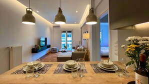 A modern dining area with a wooden table set for four, overhead pendant lighting, and a view into a living space with a sofa and a TV. - AgiasFoteinis20  ·  Nea Smirni Square chic apartme (Athens)