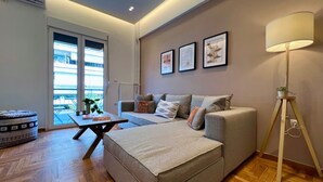 Een moderne woonkamer met een bank, een salontafel en een vloerlamp. Er hangen fotolijstjes aan de muur en een raam met uitzicht op buiten.