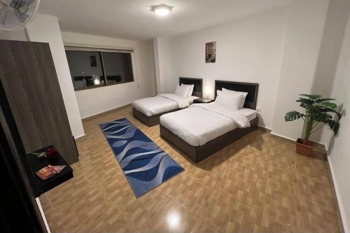A hotel room with two beds, a large window, a fan, a blue rug, and a potted plant.