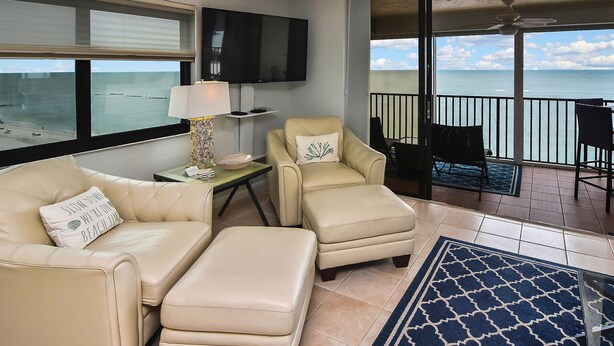 A living room with a beige sofa, two armchairs, and a TV on the wall. - SEA WINDS 1201  
Beach Front Getaway with spectacular views of the Gulf! (Marco Island)