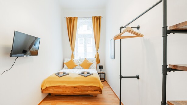 A bedroom with a wooden bed, a TV mounted on the wall, a bedside table, and a hanging clothes rack. - 3 Bedrooms - 72sqm - 15 Min. City Center (Vienna)