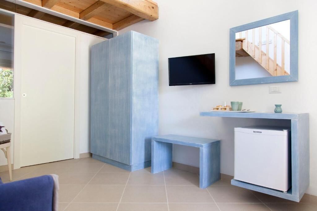 A compact room with a white door, a blue cabinet, a small bench, a mounted TV, and a white mini-fridge.