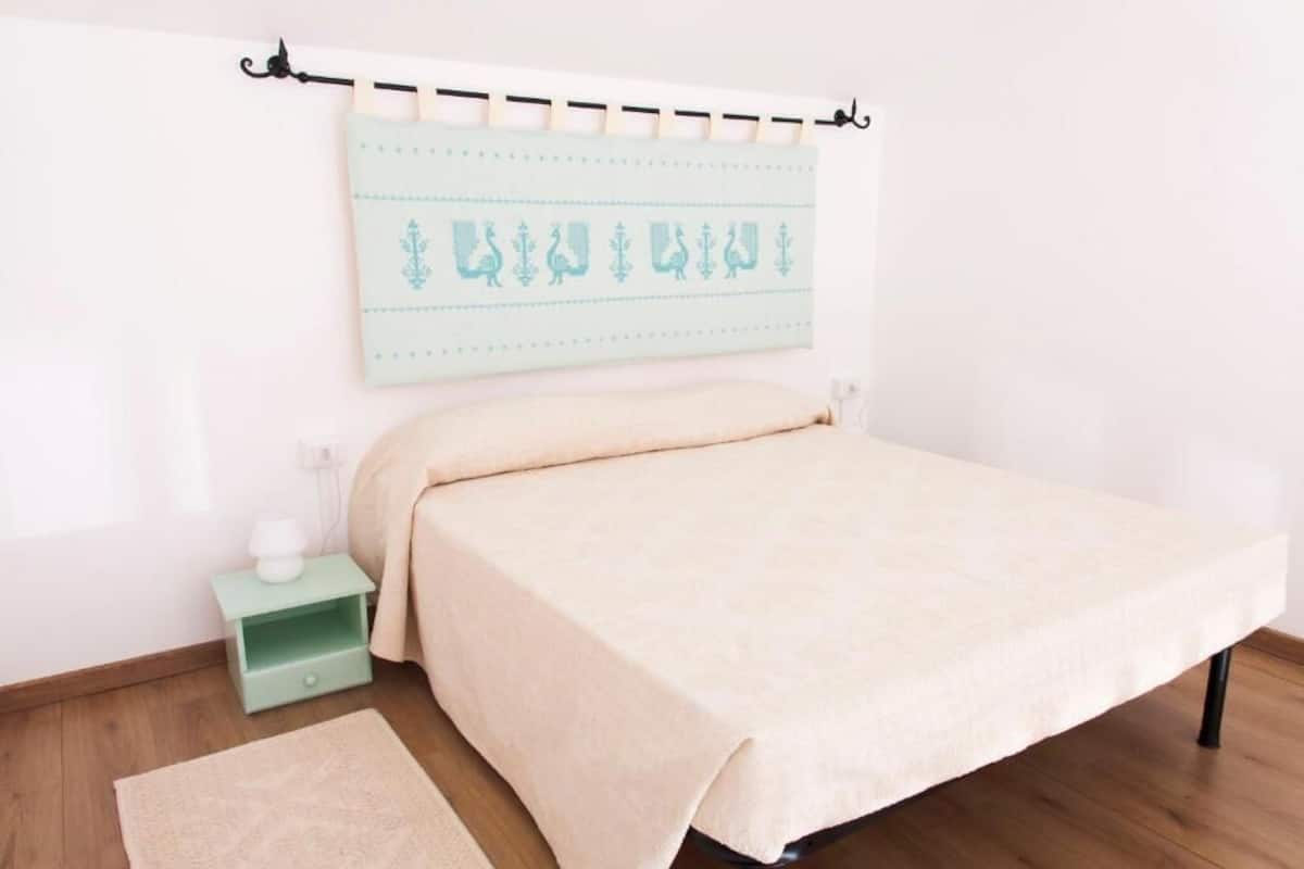 Een net opgemaakt bed met een lichtgekleurd sprei, een klein nachtkastje met een lamp, en een wandbeugel voor een gordijnroede met een lichtblauwe, geribbelde gordijn.