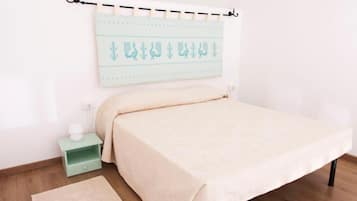 Een net opgemaakt bed met een lichtgekleurd sprei, een klein nachtkastje met een lamp, en een wandbeugel voor een gordijnroede met een lichtblauwe, geribbelde gordijn.