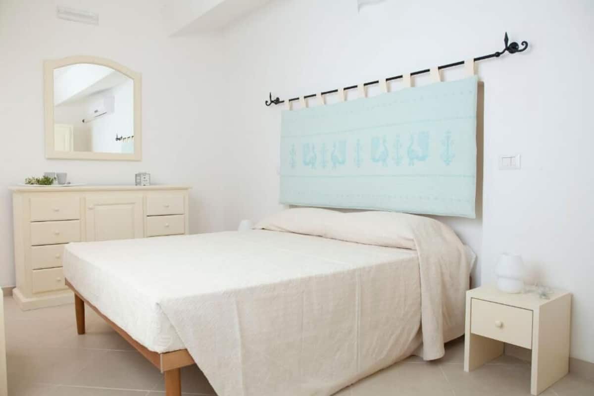 Een slaapkamer met een bed, nachtkastje, dressoir, spiegel en een kunstwerk aan de muur.