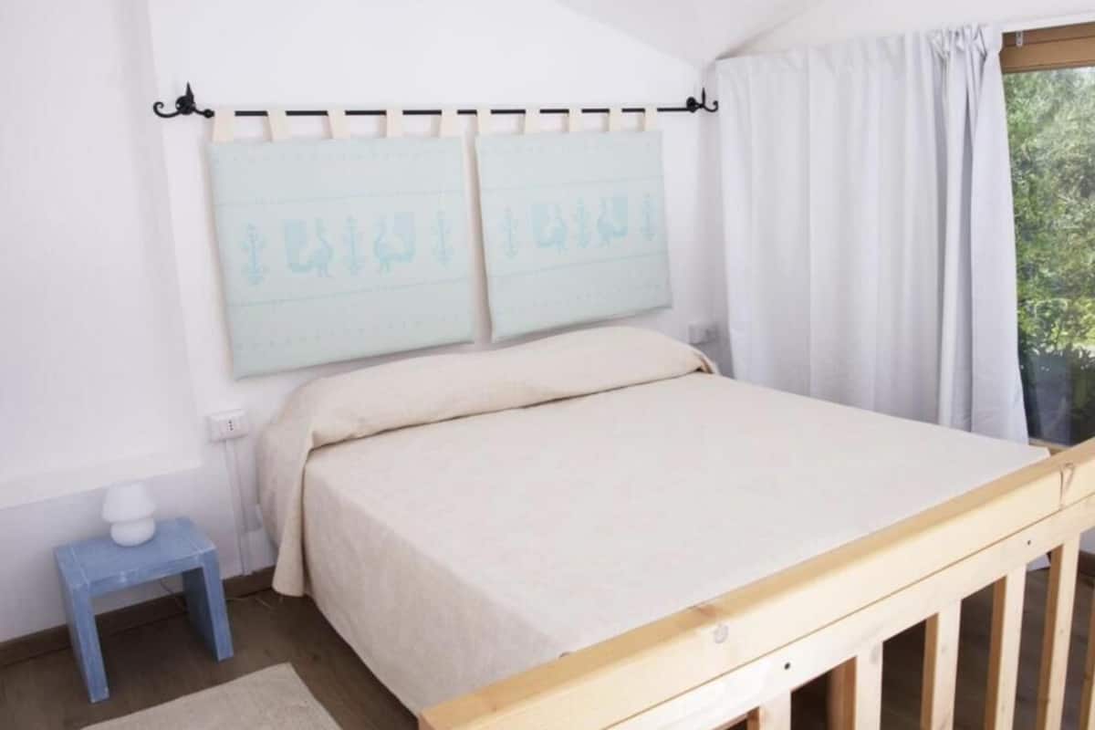 Een slaapkamer met een bed, een klein nachtkastje, een raam met gordijnen en een wandlamp.