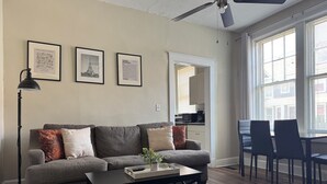 Ein Wohnzimmer mit grauem Sofa, einem schwarzen Couchtisch und einem gemusterten Teppich. An den Wänden hängen gerahmte Bilder, eine Deckenlampe und durch eine offene Tür ist die Küche sichtbar.