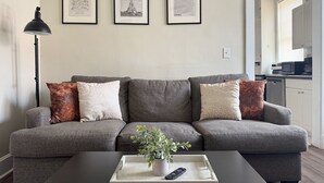 Ein Wohnzimmer mit grauem Sofa, drei gerahmten Bildern an der Wand, einer Stehlampe und einem Couchtisch mit einer Auflage, auf der sich eine Pflanze und eine Fernbedienung befinden.