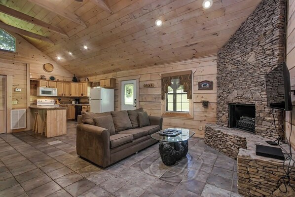 A cozy cabin interior with a stone fireplace, wooden ceiling, and a kitchenette.