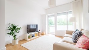 A modern living room with a flat-screen TV, a white sofa, a potted plant, and a large window with curtains.
