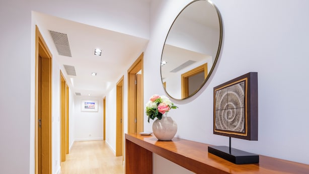 A hallway with wooden flooring, a reception desk with a vase of flowers, a round mirror, and a framed artwork.