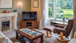A living room with a fireplace, television, comfortable seating, and a view of greenery. - 3 bedroom accommodation in Keswick (Keswick)