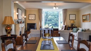 A dining area with a wooden table, chairs, a fireplace, and a view of greenery through large windows. - 3 bedroom accommodation in Keswick (Keswick)