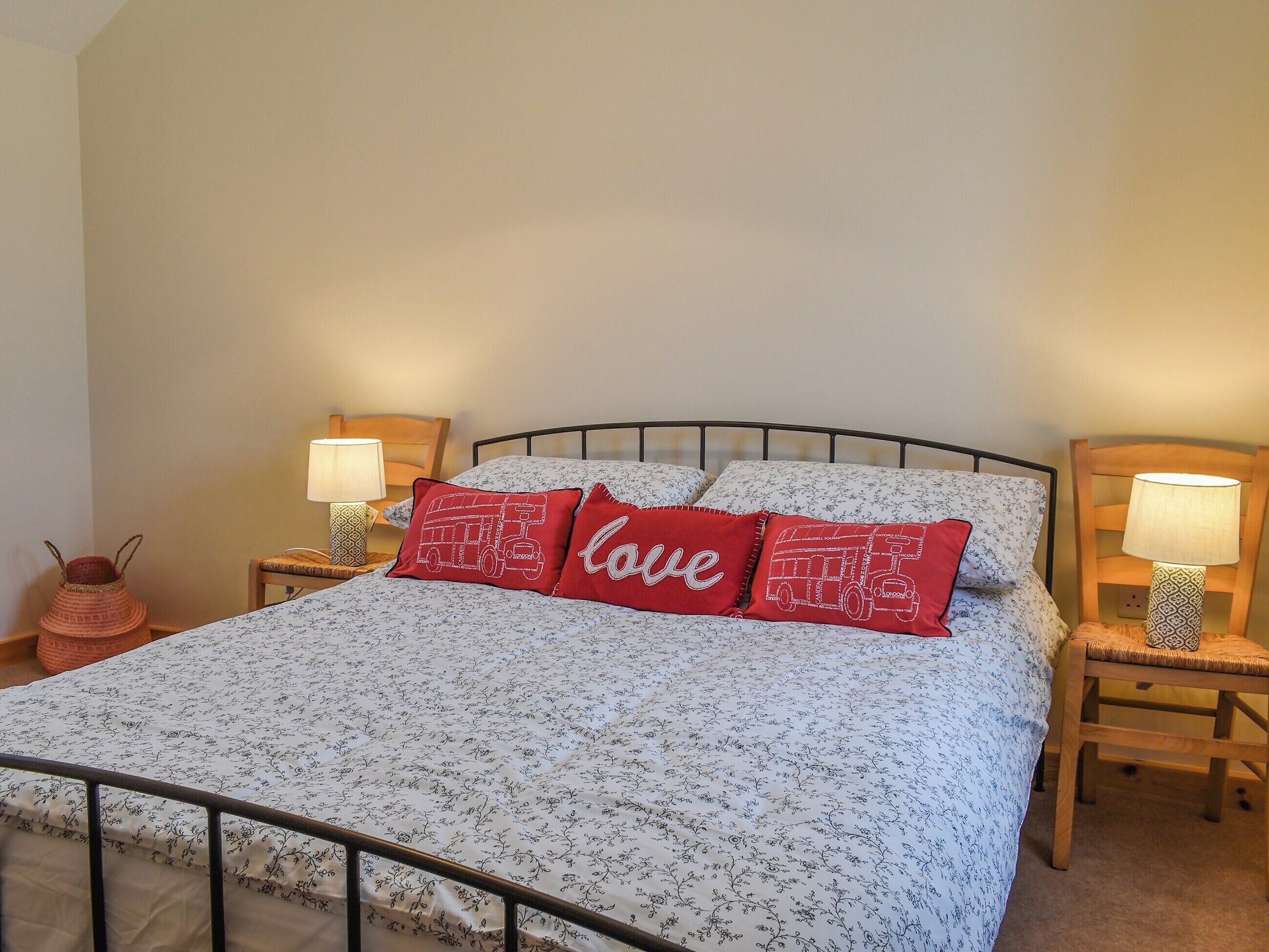 Een netjes opgemaakt bed met bloemenbeddengoed en twee rode kussens met de tekst 'love'. Twee nachtkastjes met lampen en een kleine potplant staan ernaast.