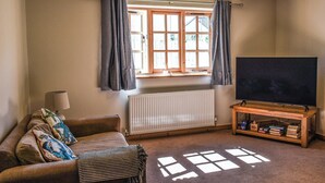 A cozy living room with a sofa, a television on a wooden stand, a radiator, and a window with curtains. - 2 bedroom accommodation in Par, near Fowey (Par, near Fowey)