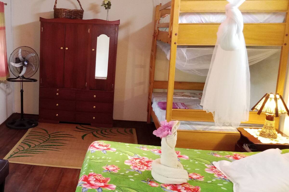 A room with a wooden bunk bed, a desk with a lamp, a wardrobe, a fan, and a floral tablecloth.