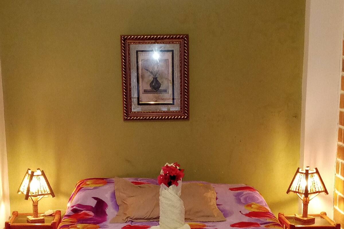 A bedroom with a bed covered in a floral bedspread, two bedside lamps, a framed picture on the wall, and a window above the bed.