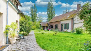 A courtyard with a stone pathway, a small table, and chairs. There are potted plants and a grassy area with flowers.